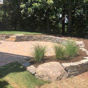 Outdoor patio with stone retaining wall, ornamental grasses, large rocks, and trees in background