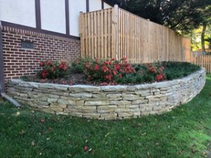 Raised stone garden bed with red flowers and shrubs along brick building and wooden fence.
