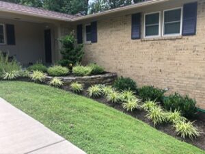 Brick house side with landscaped garden bed featuring ornamental grasses, shrubs, stone borders, and sidewalk.