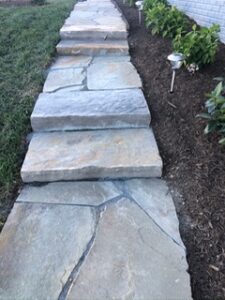 Stone walkway with uneven slabs bordered by grass, garden bed with plants, solar light, and brick wall.
