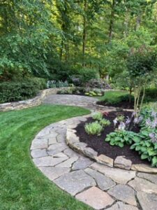Curved stone pathway dividing grass and flower beds in a garden with trees and shrubs.