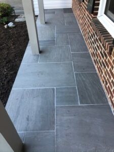 Gray stone tile patio beside a brick wall with white posts and a small garden area.