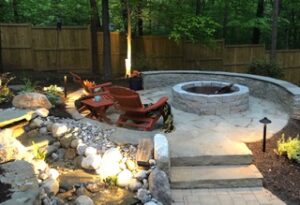 Outdoor stone patio with circular fire pit, curved stone bench, wooden chairs, and surrounding fence.