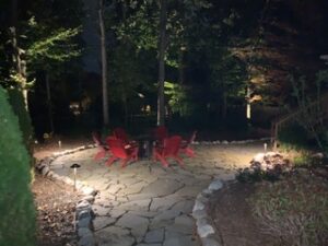 Nighttime stone patio with red chairs around a fire pit, illuminated by ground lights.