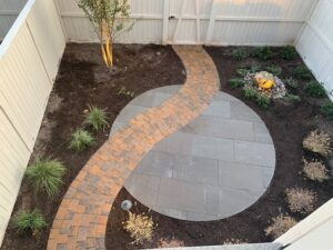 Backyard with curved brick path, circular stone patio, small plants, and lit fire feature.
