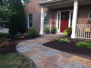 Brick house with covered porch, red door with wreath, stone walkway, and landscaped plants.