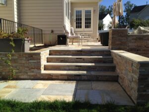 Stone steps lead from a tiled patio with stone retaining walls to a raised deck with seating.