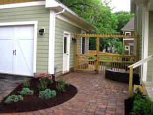 Paved patio with garden bed, stone fire pit, wooden pergola, and light green house exterior.