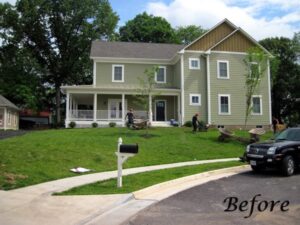 Two-story green house with porch, workers landscaping sloped front yard, black car parked nearby.