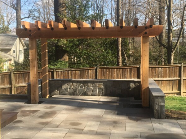 Wooden pergola on stone tile patio with stone wall and wooden fence in backyard.
