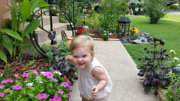 Toddlers wearing a white sleeveless top on a garden pathway with flowers and concrete steps.