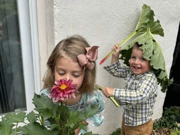 Child smelling pink flower while another child holds large green leaves outdoors by a wall